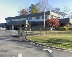 Office building with a fenced parking lot, surrounded by autumn trees. A few cars are parked, and there's a sign near the entrance.