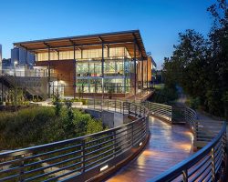 Commercial Projects Modern building with large glass windows, surrounded by trees, and a curved walkway at dusk. Industrial tanks visible in the background.