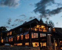 Design-Build Projects A large, rustic lodge building with illuminated windows, surrounded by trees, under a partly cloudy evening sky.
