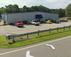 A small industrial building with parked cars in front, located near a road with a metal guardrail. A sign reads "Horsepower Motorsports.