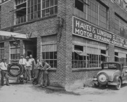 Vintage photo of men outside Hayes & Lunsford Electric Shop, with a car parked in front and a large tire being moved.