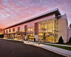 Healthcare Projects A modern two-story building with glass windows, surrounded by a paved area, under a colorful sunset sky.