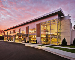 Healthcare Projects A modern two-story building with glass windows, surrounded by a paved area, under a colorful sunset sky.