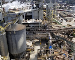 Industrial Projects Aerial view of an industrial plant with large storage tanks, pipes, and chimneys emitting smoke under a clear blue sky.